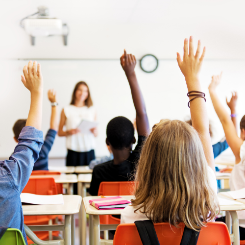 A number of hands up in a classroom with the teacher standing at the front of the class out of focus.
